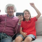 Bukan Salah Gen Z: Membongkar Mitos Konflik Antar-generasi Lewat Kacamata Sejarah Smiling grandfather and granddaughter enjoying media on a smartphone at home.